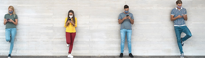 Young people wearing surgical masks, using their smartphones while waiting in line and socially distancing.